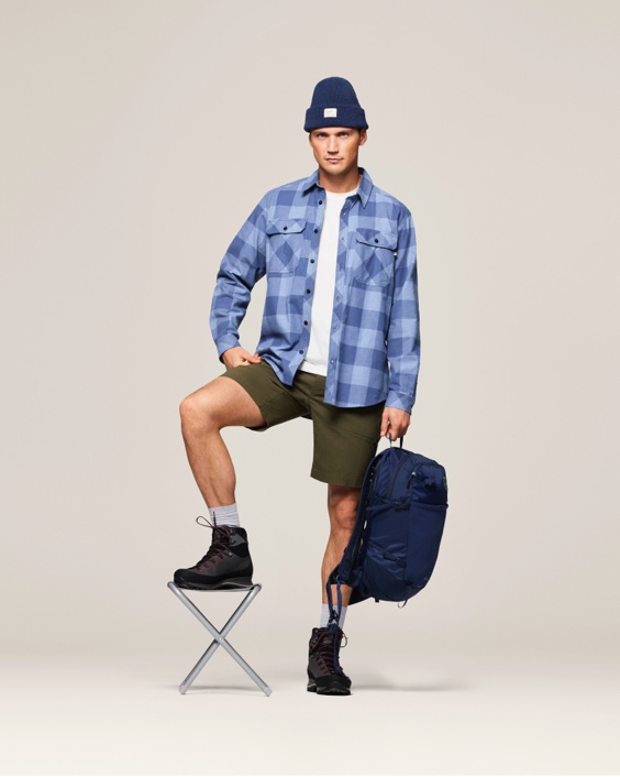 Studio shot of a man posing in a blue plaid button-down shirt, beanie, shorts, and boots while resting a foot on a small stool.