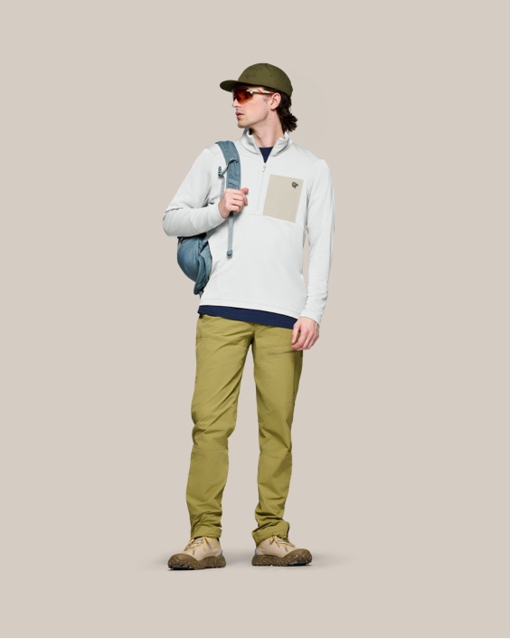 Studio shot of a man modeling a grey fleece pullover, green pants, and a cap.