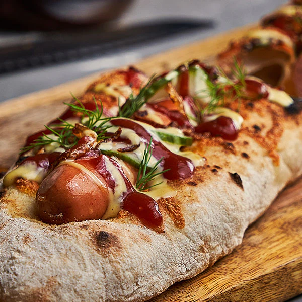A hot dog nestled into a pizza dough crust topped with sauces and pickles