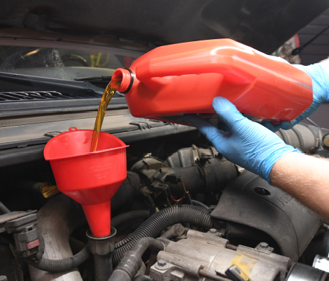 A red jug of yellow motor oil held by hands with blue gloves being poured via plastic red funnel into a vehicle's engine.