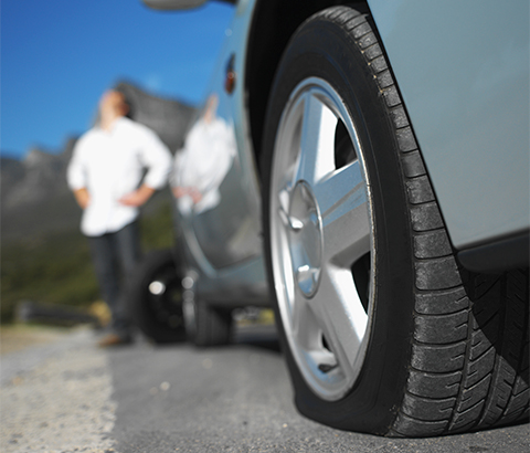 Close-up of a vehicle's flat tire with a person in a white shirt holding their hands on their hips and staring upward unfocused in the background.