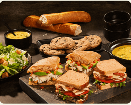 A table laden with a variety of food items, including three sandwiches cut in half, a bowl of green salad with croutons, two bowls of yellow soup, several chocolate chip cookies, and two baguettes. The sandwiches feature different fillings and breads, showcasing fresh ingredients.