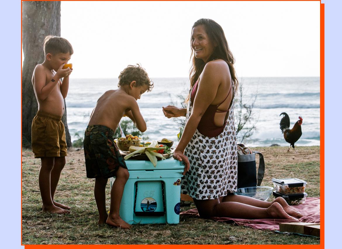 Two kids and an adult eating food off the top of a cooler at the beach.  