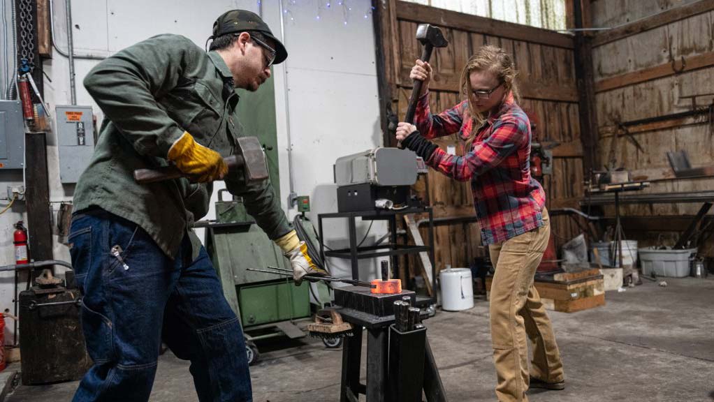 Two blacksmiths taking turns hammering a glowing hot chunk of metal.