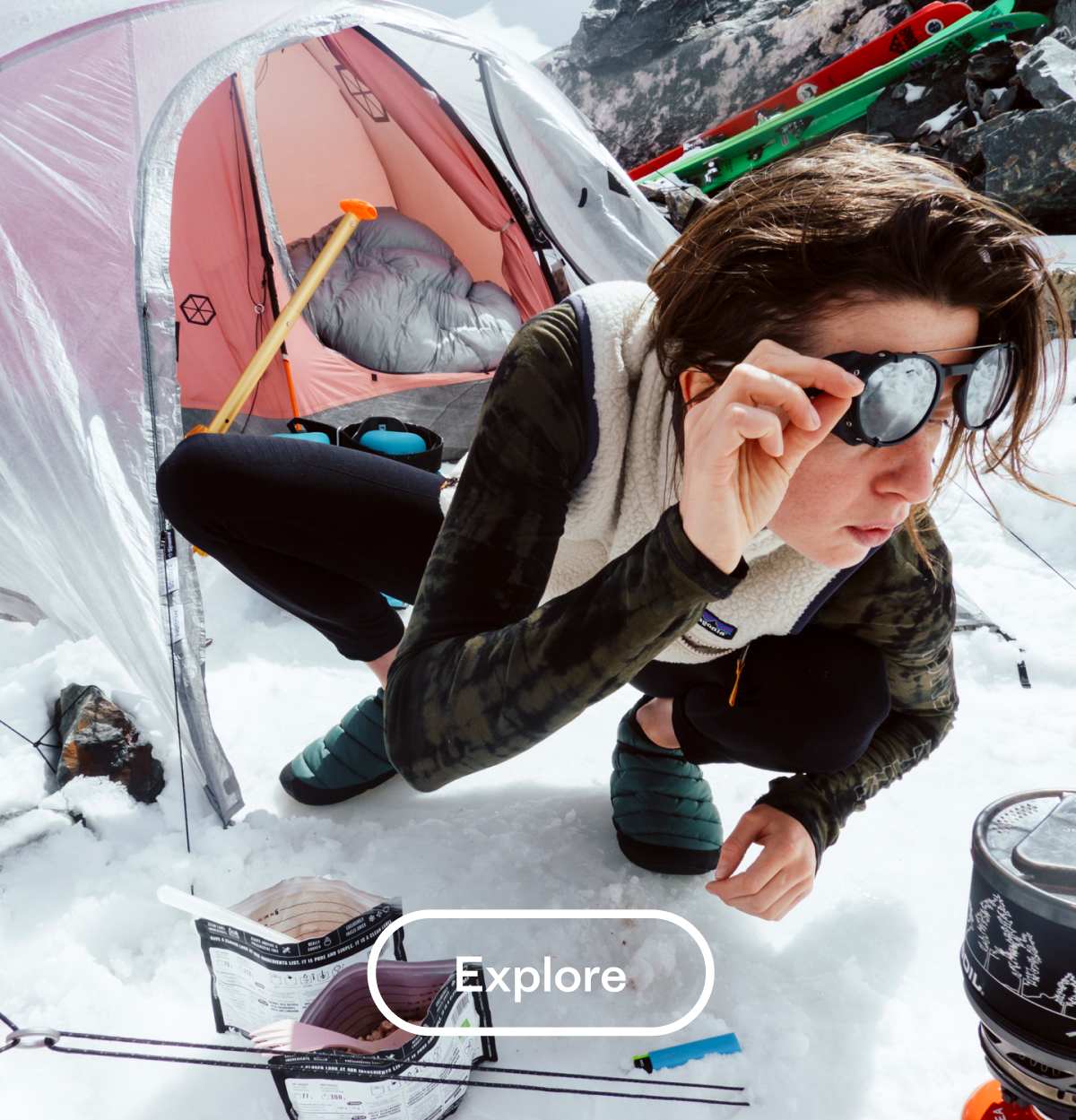 A backcountry skier boils water on her stove in front of her tent on the snow.