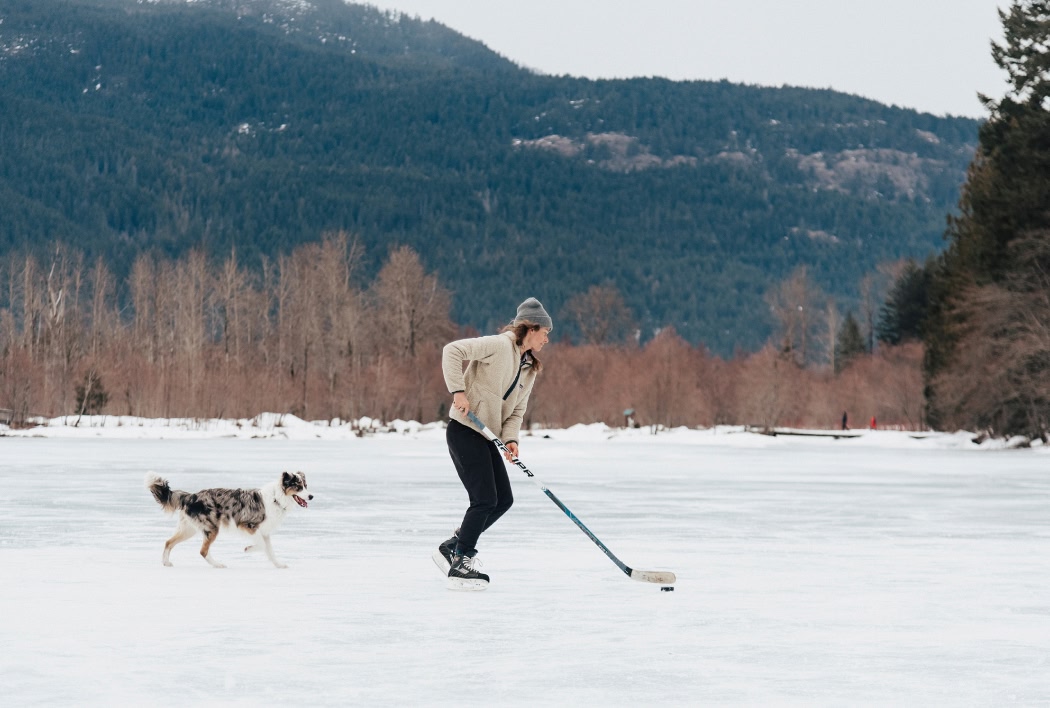 A person ice skates with a hockey stick and a dog following.