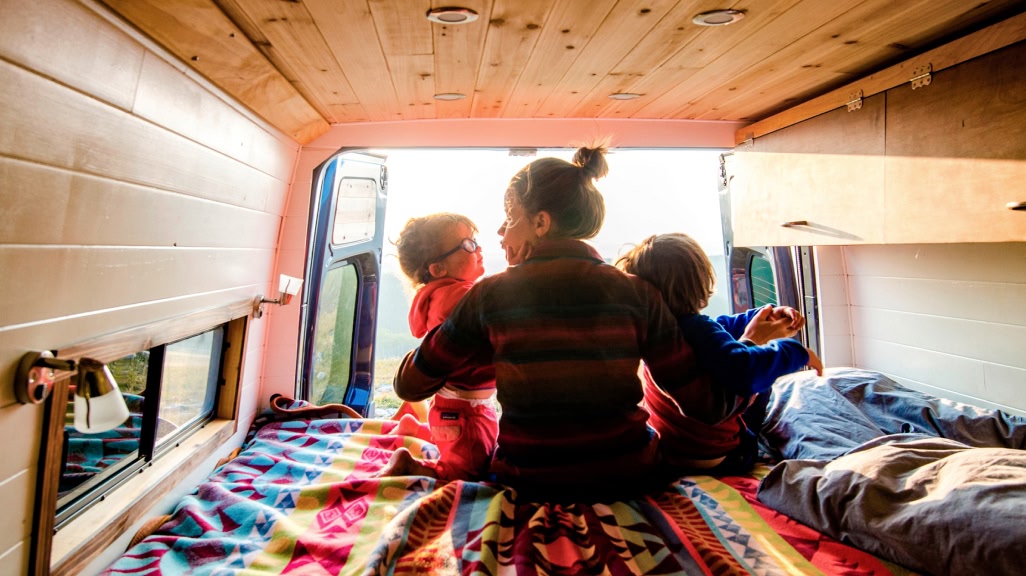 A mom and two kids sit on the edge of a bed in the back of a van.