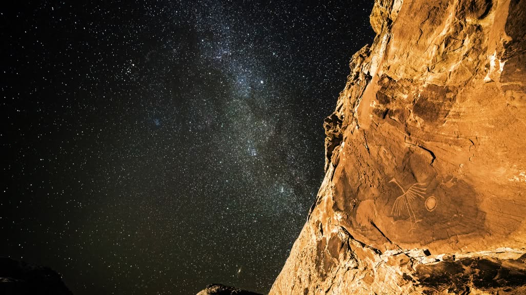 Stars in the night sky shine brightly above a petroglyph on a rock wall.