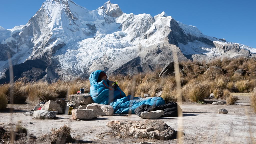 A person leans against a rock while wrapped in a mummy sleeping bag with a snowy peak in the background.