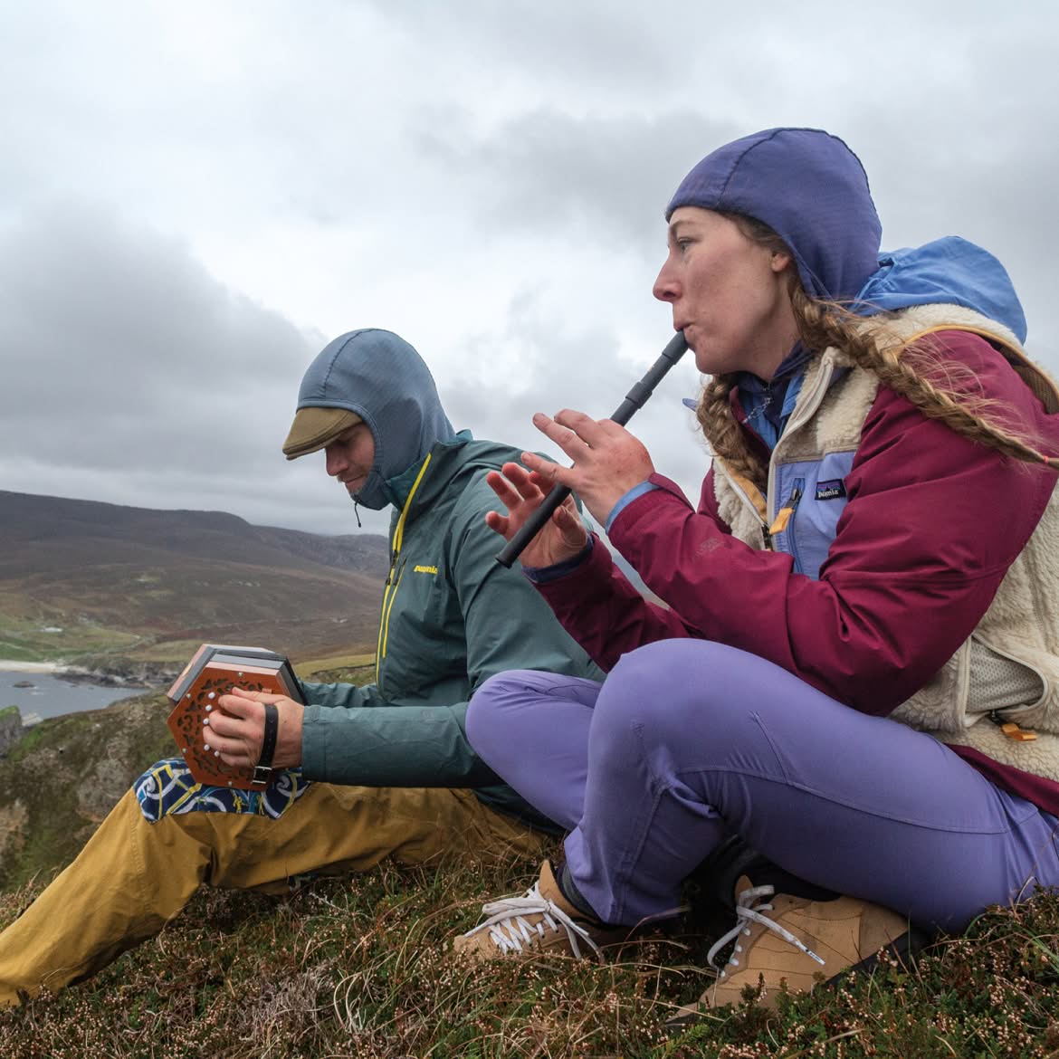 Two people play musical instruments while sitting outdoors on a cold, cloudy day.