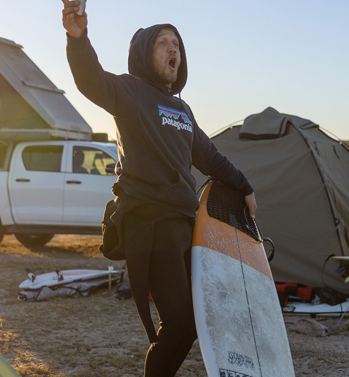 A person stands in campsite holding a surfboard in one hand and a bar of wax in the other.