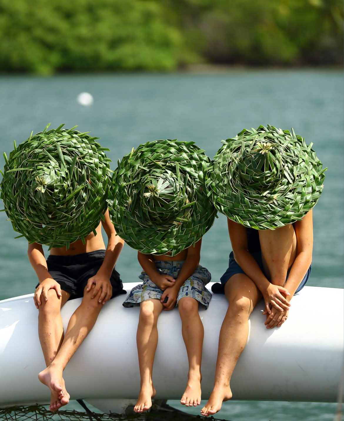 Three people sit alongside each other by a body of water wearing woven sun hats that hide their faces.