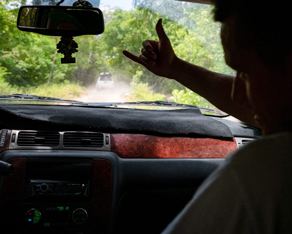 A photo taken from the backseat of a car following another car down a  dirt road. We see the driver’s hand on the wheel and the left side of the passenger’s  face. Passenger is making a “shaka” hand sign. 
