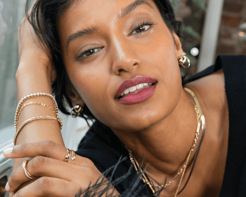Woman wearing layered gold necklaces, gold hoop earrings, and stacked gold bracelets and rings, posing with her hand near her face.