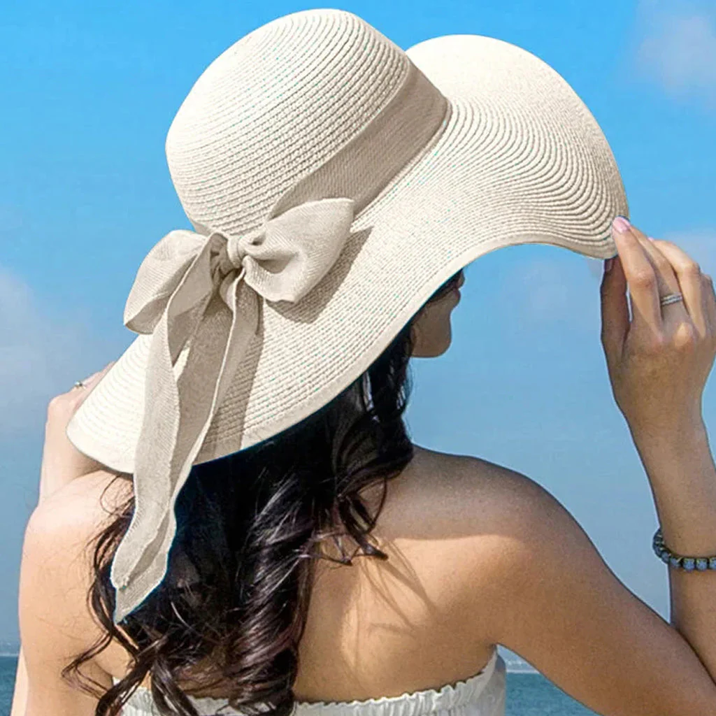 Woman wearing oversized cream straw sun hat with bow tie against blue sky and ocean background