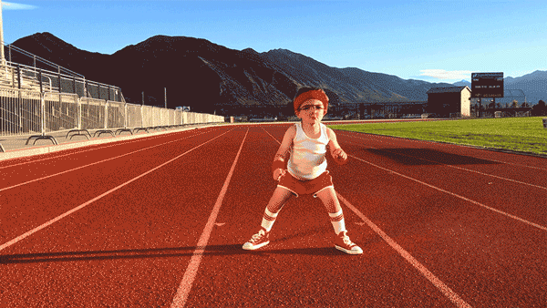 A smiling six year old boy wearing 1980s workout clothes, including a red sweatband and arm bands, bounces on a red running track. In the background is a clear bright sky against a large mountain range.
