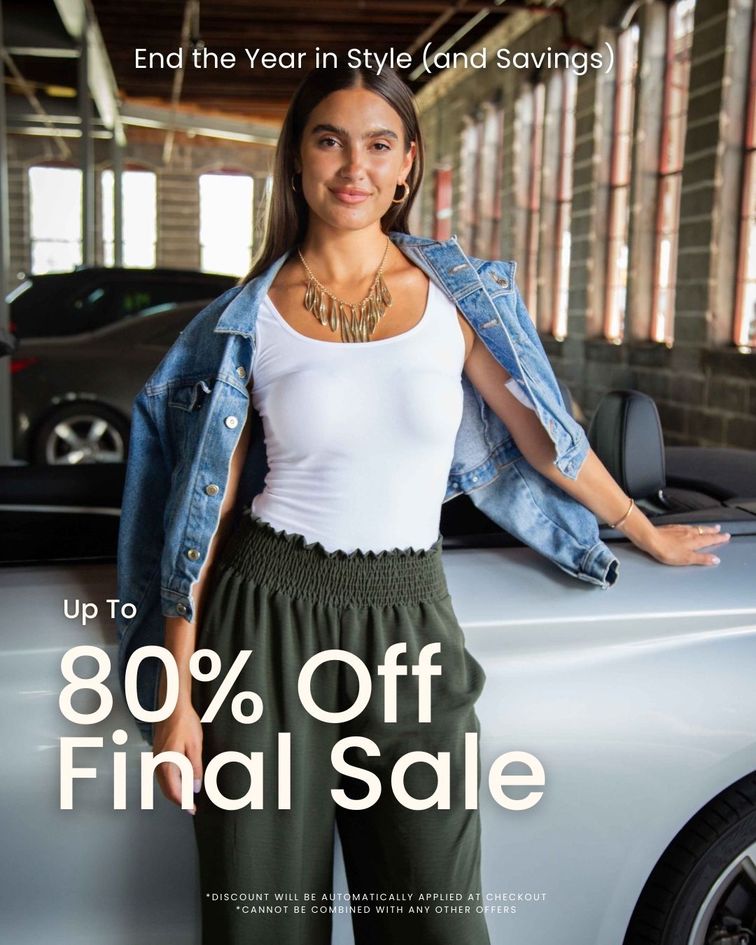 A woman standing confidently beside a silver convertible inside an industrial-style garage. She’s wearing a white tank top, olive green smocked-waist pants, a denim jacket draped over her shoulders, and a gold statement necklace. The promotional text on the image reads “End the Year in Style (and Savings)” and “Up to 80% Off Final Sale.