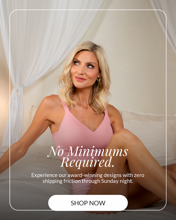 Blonde woman sitting comfortably on a bed in a soft, neutral-toned bedroom, wearing a pink seamless bra and smiling off to the side. Overlay text reads “No Minimums Required. Experience our award-winning designs with zero shipping friction through Sunday night.” A “Shop Now” button appears at the bottom.