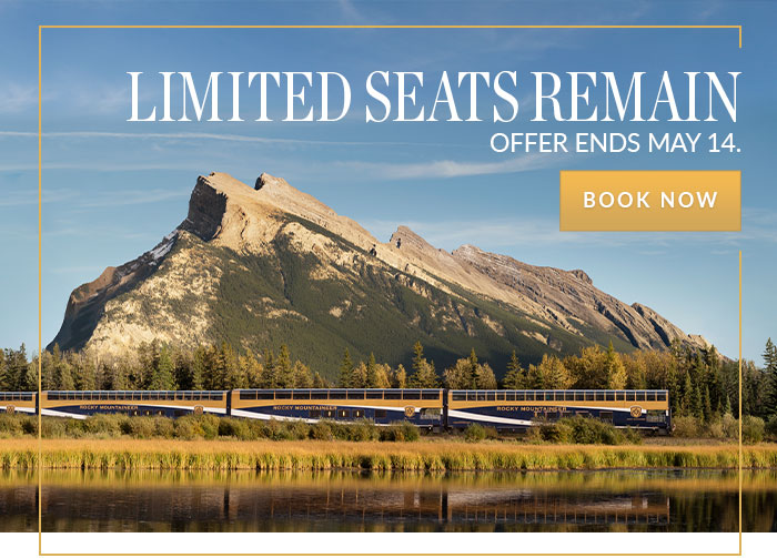 A Rocky Mountaineer train passing a lake with forest and mountains in the background.