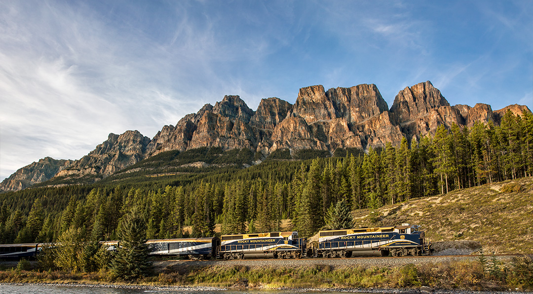 Rocky Mountaineer train passing a river with mountains in the background.