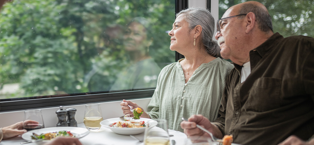 Guests enjoy a meal aboard a Rocky Mountaineer train while looking out at the passing scenery through large windows.