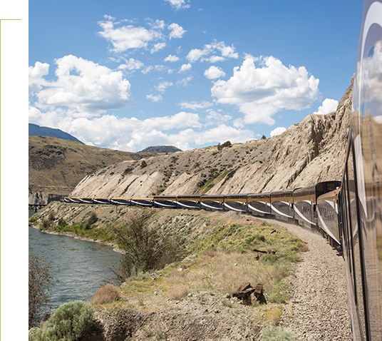 View from a Rocky Mountaineer train winding along a river through a rugged canyon landscape under a bright blue sky.