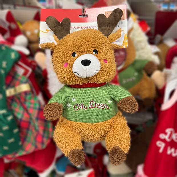 Plush reindeer dog toy wearing a green sweater with the words “Oh Deer” embroidered in white and red, displayed on a store shelf among other holiday-themed pet toys.
