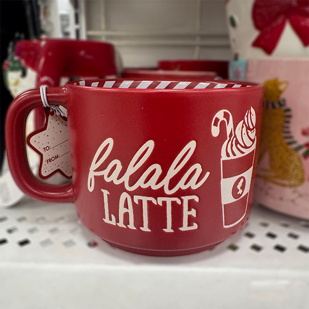 A red ceramic mug with white lettering that reads “falala LATTE,” featuring a graphic of a holiday drink with a candy cane and whipped cream. 