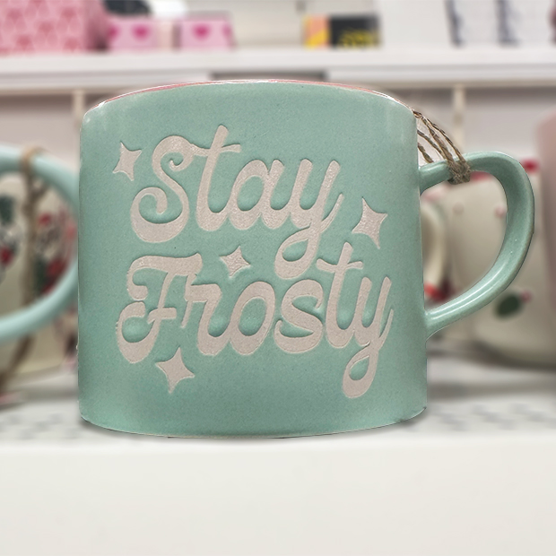 A mint green ceramic mug with white lettering that reads “Stay Frosty,” decorated with star accents. 
