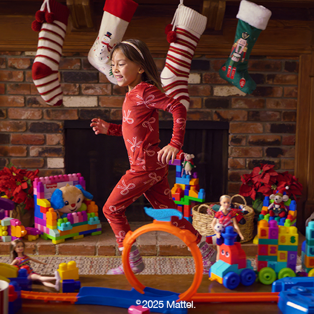 Child in red holiday pajamas playing with colorful building blocks and toy trains in front of a decorated fireplace with stockings and holiday decorations. 