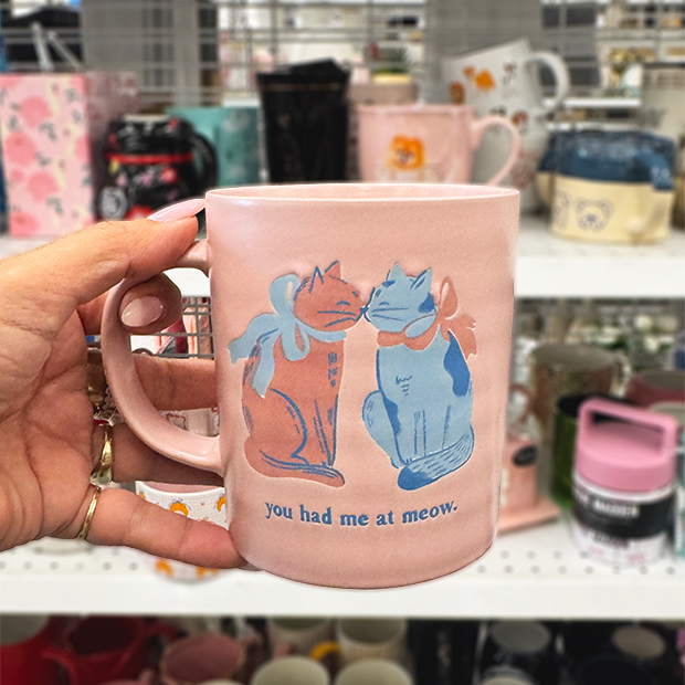 A hand holds a pink ceramic mug featuring two illustrated cats with bows touching noses, with the text “you had me at meow” beneath them. 