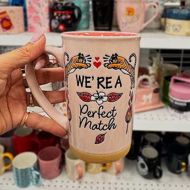 A hand holds a tall light-pink mug featuring illustrations of two tigers, small hearts, flowers, and leaves, along with the text “We’re a Perfect Match.” 