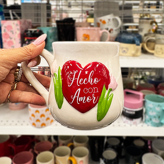 A hand holds a cream-colored ceramic mug with a large red heart and green leaves on the front, displaying the text “Hecho con Amor.” Other mugs on store shelves are visible in the background.