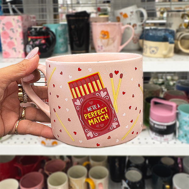 A hand holds a pink ceramic mug decorated with tiny red and white hearts and an illustration of a matchbox with matches. The matchbox reads “We’re a Perfect Match.” Store shelves of mugs appear blurred behind it.