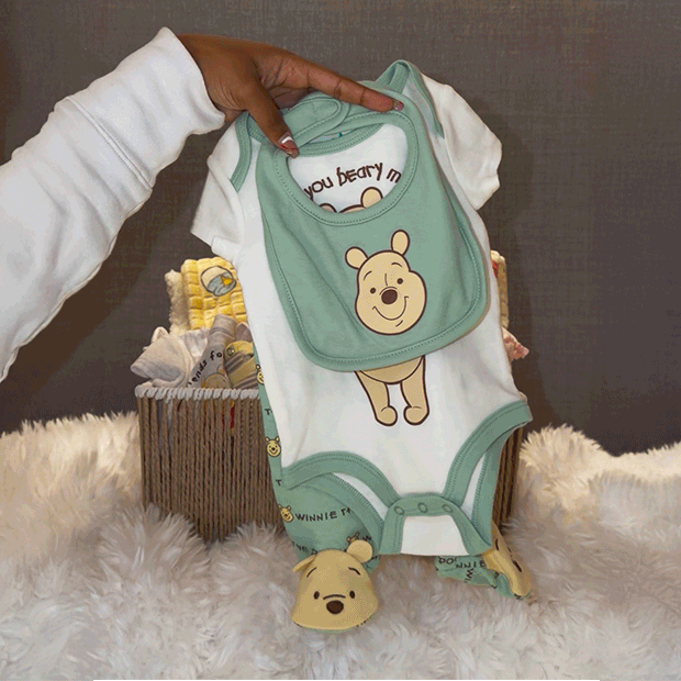 Baby outfit set featuring a white onesie with a Winnie the Pooh graphic, a matching green bib, and coordinating accessories held above a gift basket.