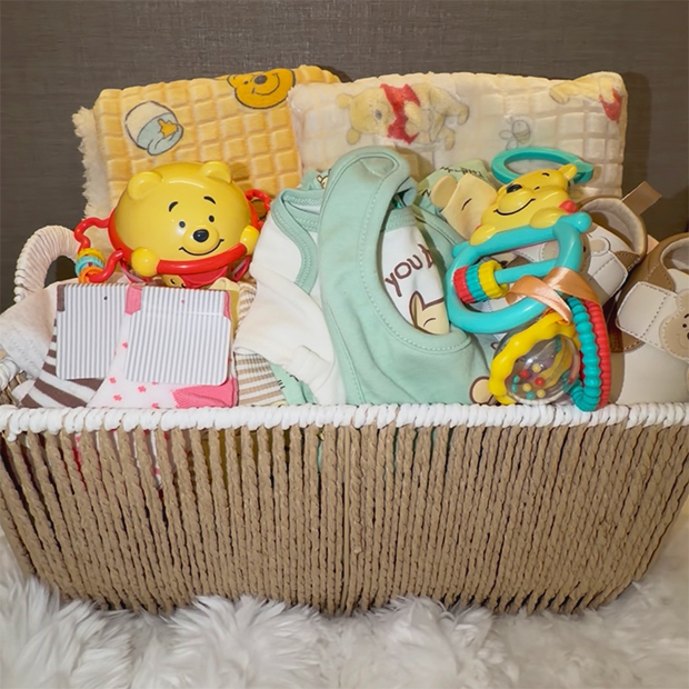 Wicker baby gift basket filled with baby essentials, including Winnie the Pooh–themed toys, a soft blanket, bibs, and small baby items arranged neatly. 