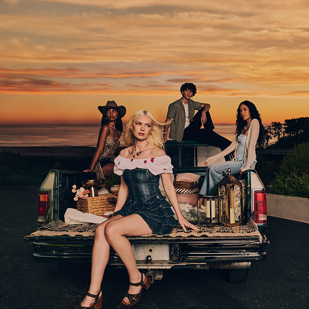 Group seated in the open tailgate of a pickup truck on a coastal road at sunset, styled with spring outfits, picnic accessories, and ocean views in the background. 