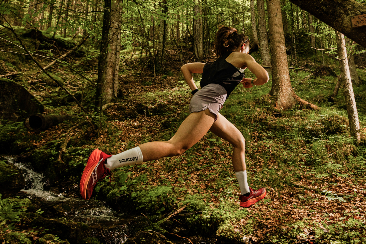 [saucony] Woman jumping over a stream in a forest wearing Saucony Peregrine shoes