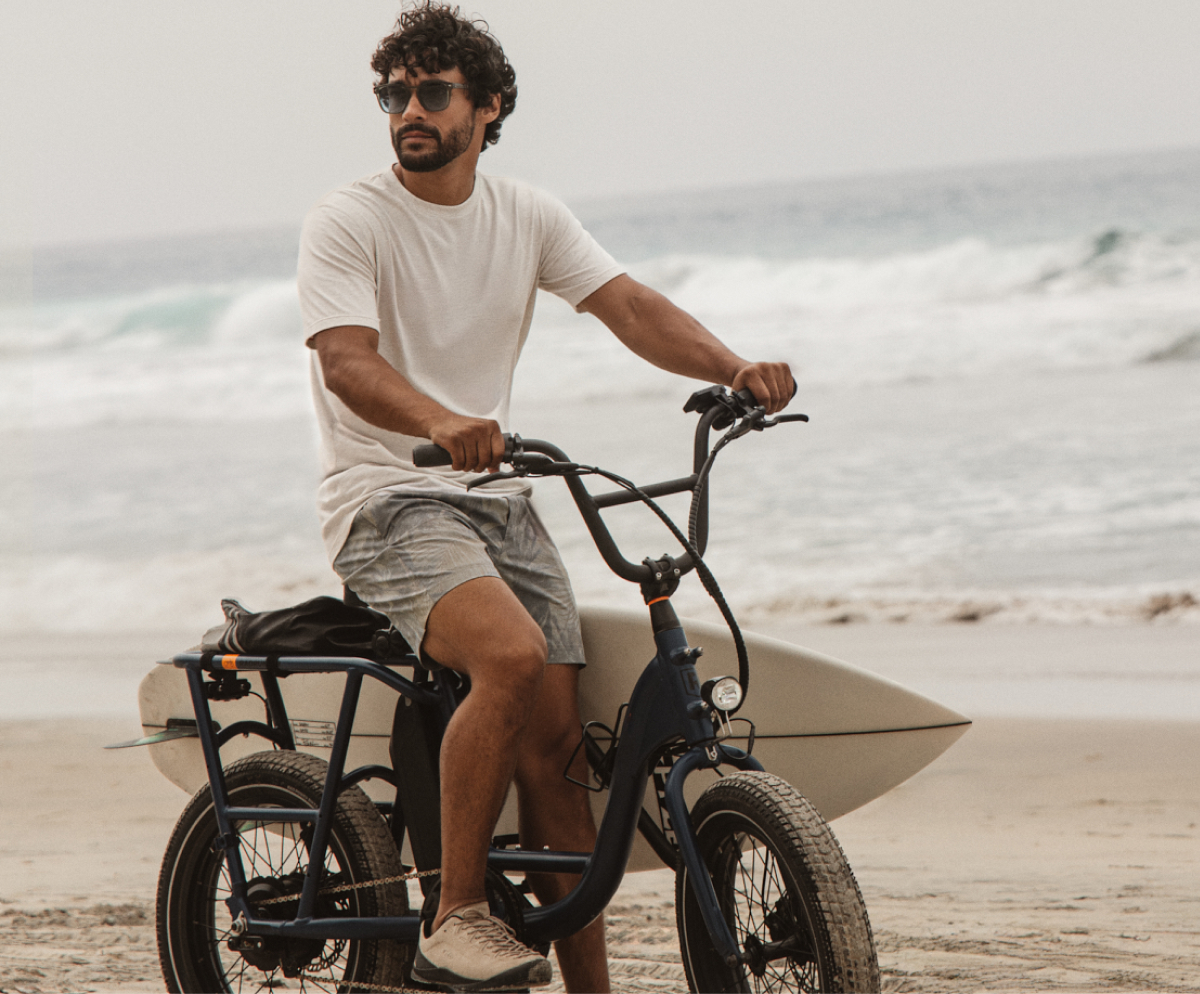 Man riding a bike on the beach with a surfboard, wearing casual coastal outfit under headline “The all-day explorer.”