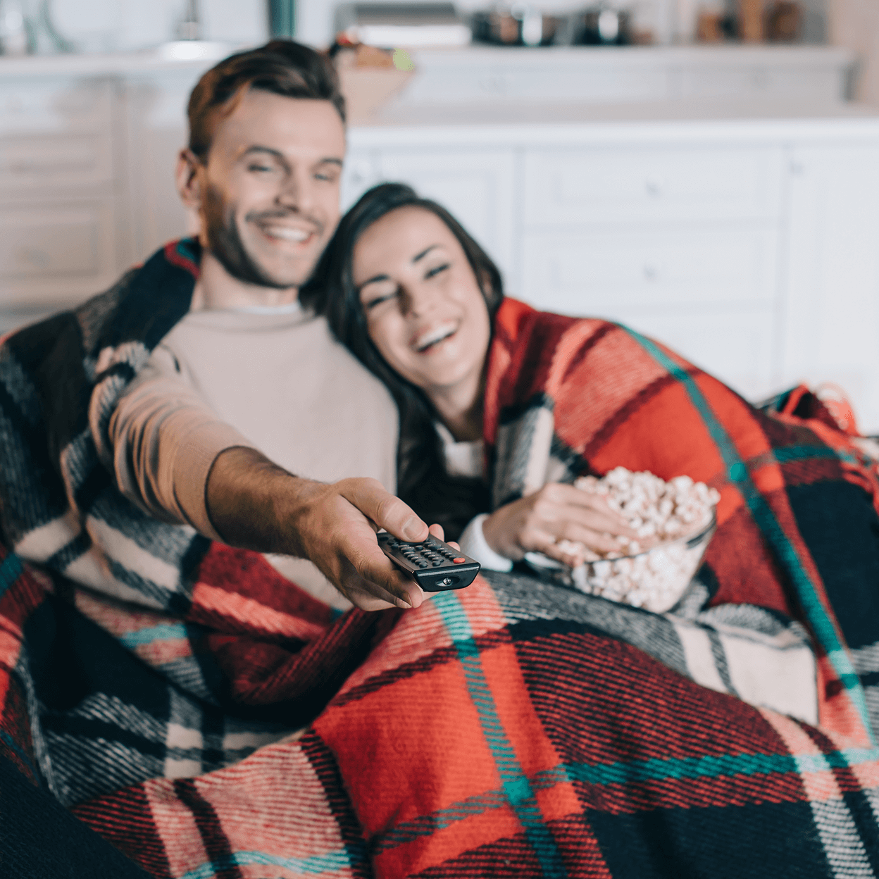 A smiling couple cuddling on a couch wrapped in a red blanket while watching TV