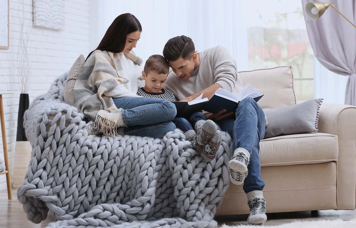 A family cuddled up on a beige couch with a grey blanket over them