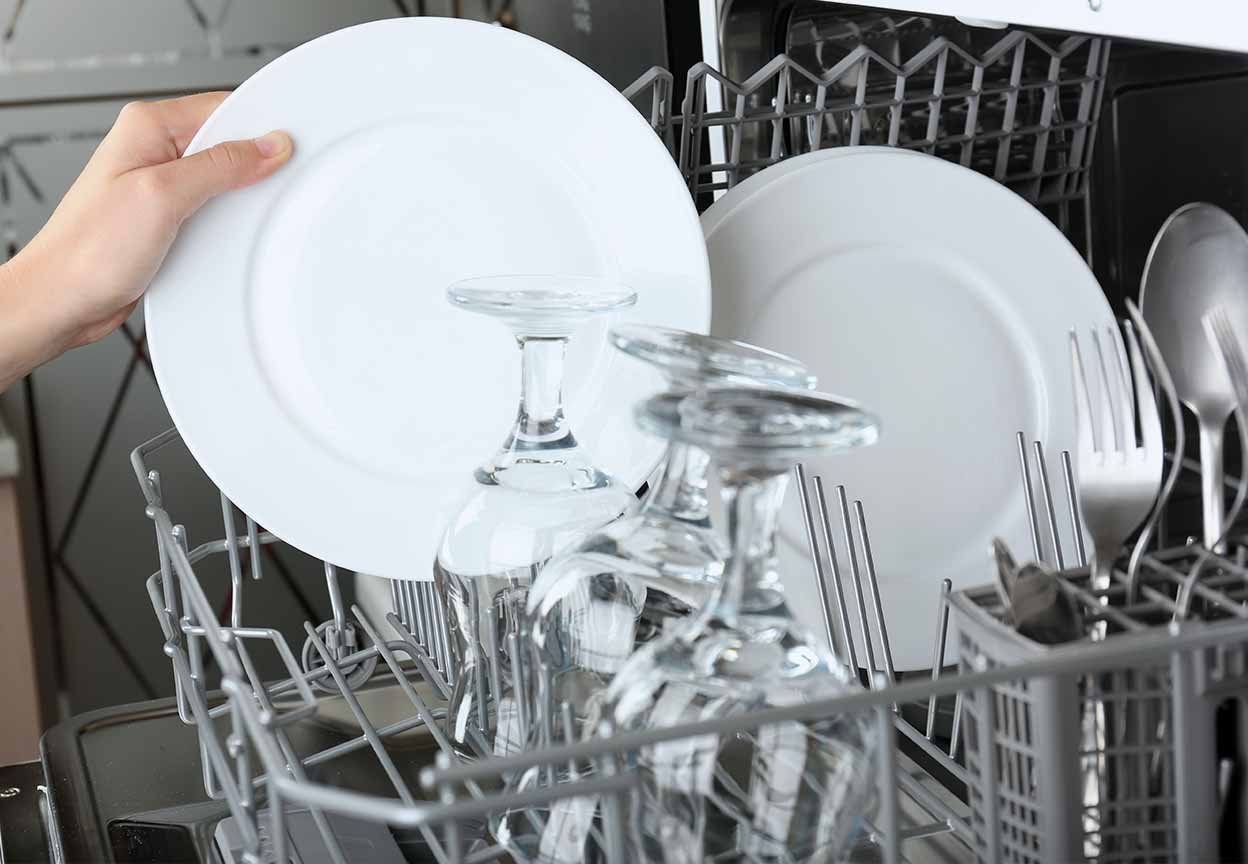 A person loading a white dish into the dishwasher