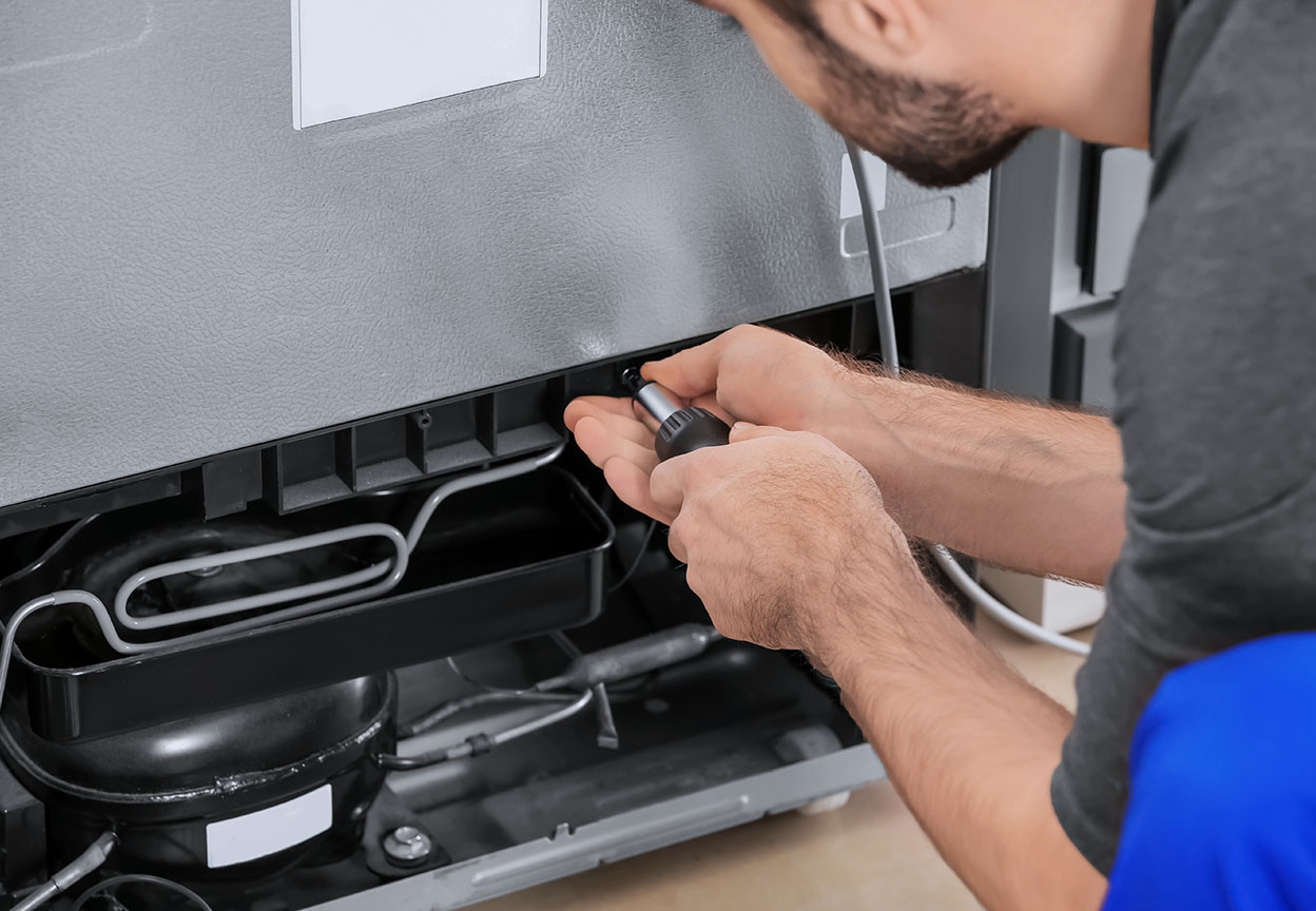 a technician kneeling in a kitchen behind a fridge unscrewing a panel