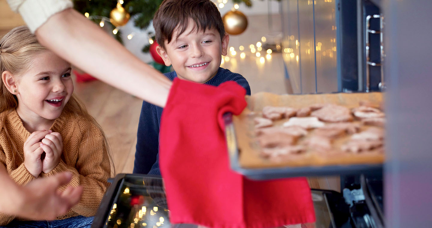 Two excited children sit by an open oven watching a tray of holiday cookies being pulled out