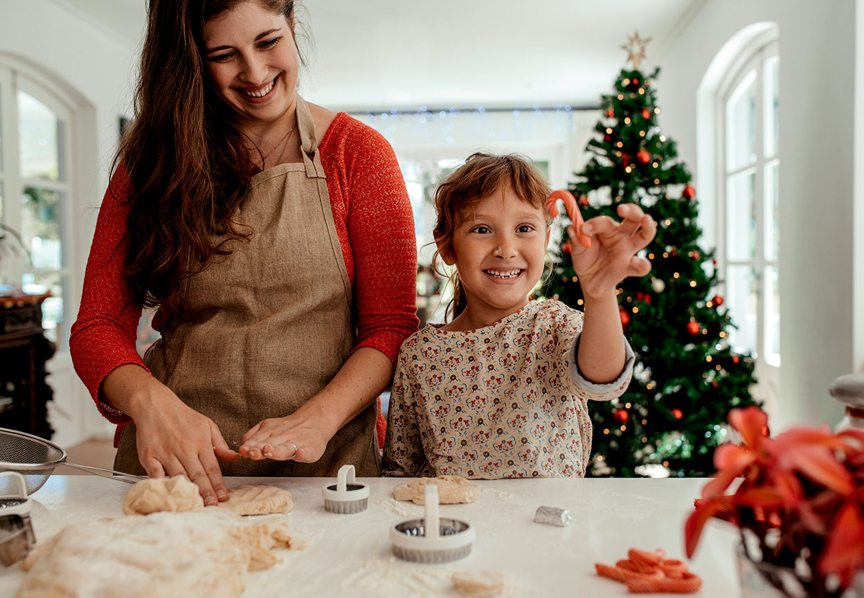 Mother and child happily shape holiday cookies at the kitchen counter while the child proudly holds a candy cane dough in front of a Christmas tree