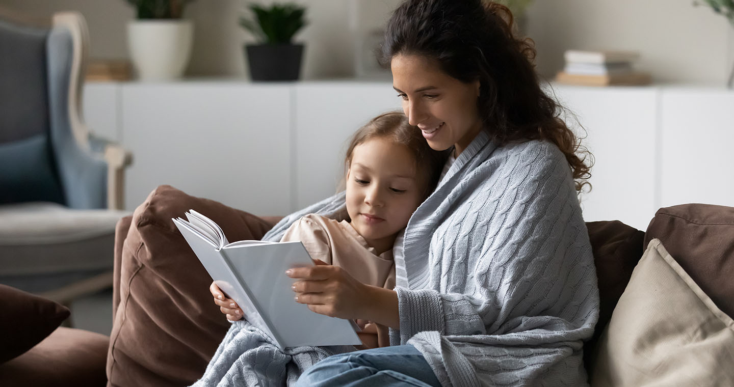 Mother and young daughter cuddle under a blanket on the couch reading a book together in a cozy living room