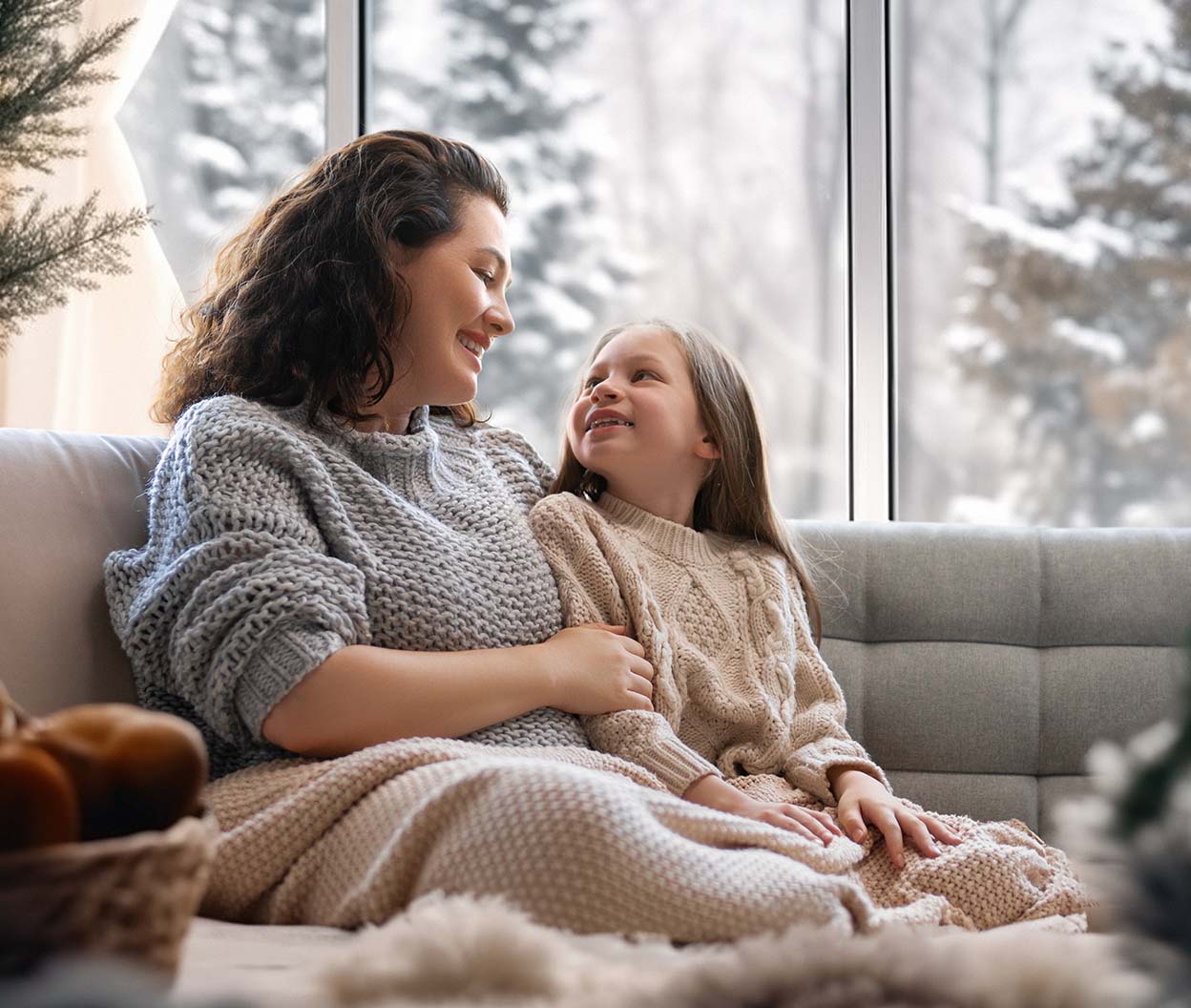 Mother and daughter relax on a cozy couch in a warm living room while snow falls outside large windows
