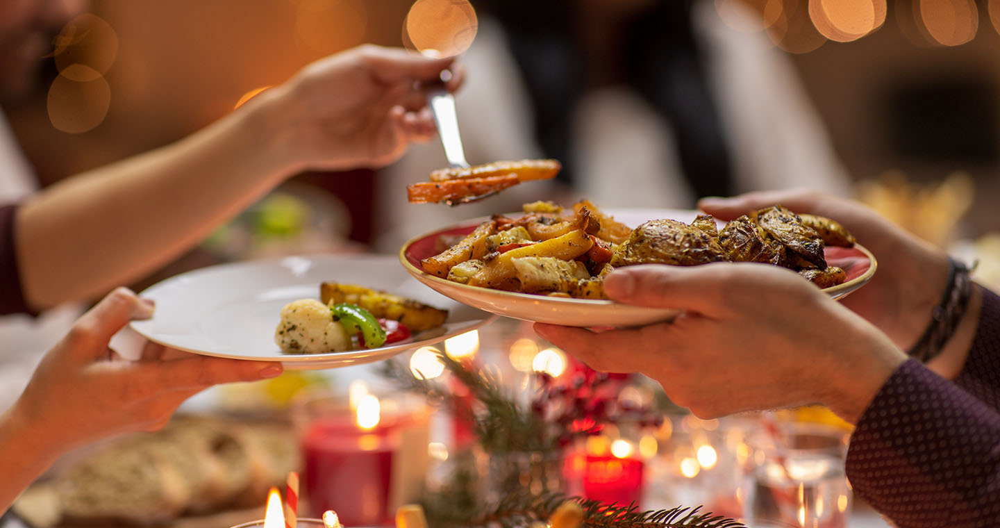 Hands serve roasted holiday vegetables from a platter at a festive candlelit dinner table