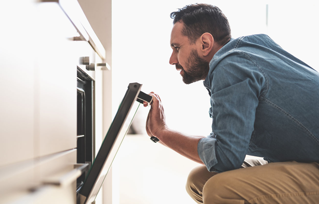 Man kneels in front of an open oven, checking inside to see how it is working