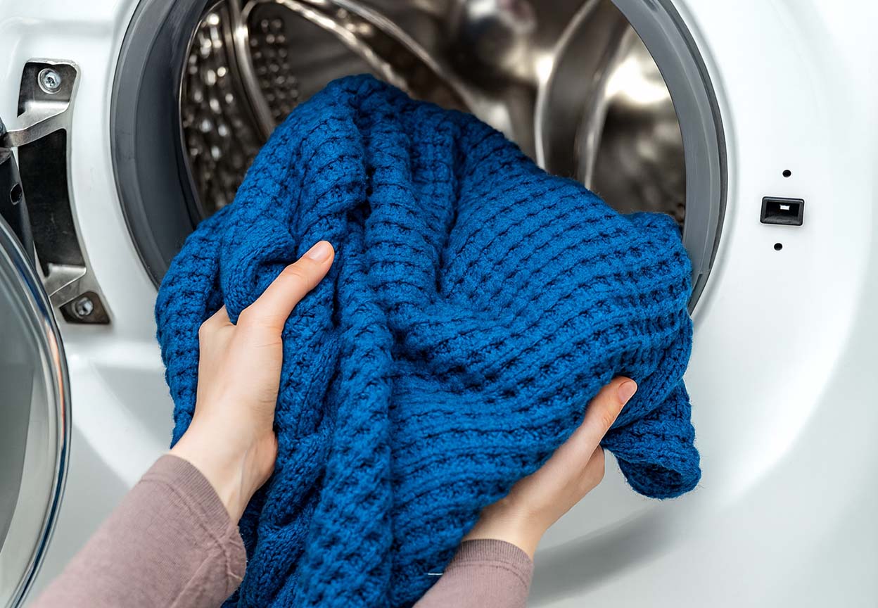 A person loads a thick blue winter blanket into a front-loading washing machine